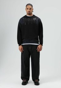 Zwarte sweatshirt met "REBELLION" in druipende witte opdruk, gecombineerd met zwarte wijde jeans en zwarte schoenen met roze veters.