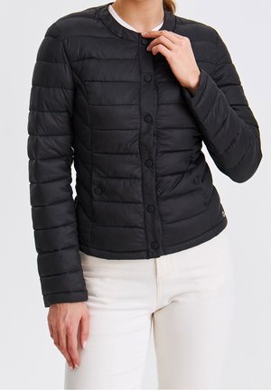 Femme portant une veste matelassée noire à boutons et un pantalon blanc, ajustant le col de la main droite.