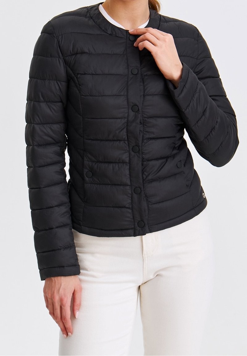 Femme portant une veste matelassée noire à boutons et un pantalon blanc, ajustant le col de la main droite.