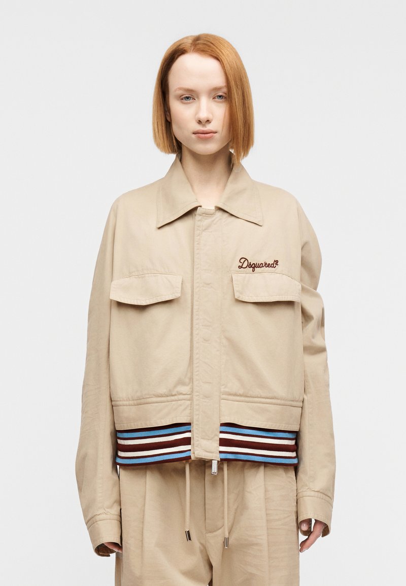 Μπεζ μπουφάν με μακριά μανίκια και δύο μπροστινέ ς τσέπες. Διαθέτει ριγωτή λουρίδα στο τελείωμα σε καφέ, μπλε και λευκό. Κεντημένο λογότυπο στο στήθος.