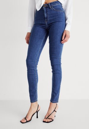 Femme portant un jean skinny bleu taille haute et des sandales noires à talons hauts et à brides, debout devant un fond uni.