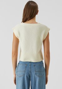 Pletený bezrukávový top v off-white barvě, s žebrovaným výstřihem a lemem, kombinovaný se světle modrými riflovými kalhotami s vysokým pasem a zadními kapsami.