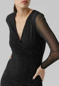 Robe noire à encolure en V, tissu scintillant, et manches longues transparentes. Coupe ajustée avec une texture lisse et un léger éclat.