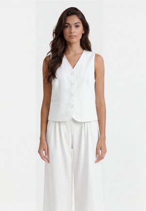 Donna con lunghi capelli scuri, indossa un gilet bianco senza maniche con bottoni e pantaloni bianchi a gamba larga, in piedi davanti a uno sfondo bianco uniforme.