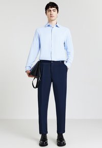 Mann trägt ein hellblaues Hemd, eine marineblaue Hose, schwarze Lederschuhe und hält eine schwarze Ledertasche vor einem einheitlichen hellen Hintergrund.