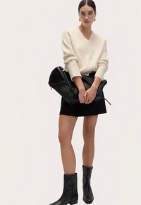 Hellbeiger, gerippter Pullover mit V-Ausschnitt, schwarzer Minirock, strukturierte schwarze Handtasche und schwarze Stiefeletten. Glatter Stoff und klare, strukturierte Form.