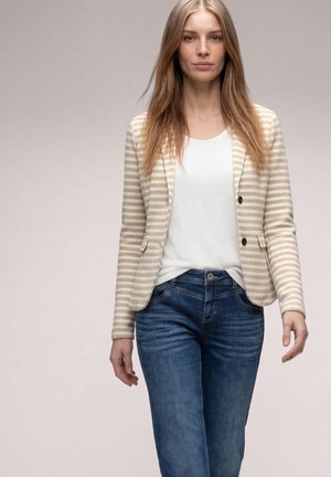 Giovane donna con capelli biondi lunghi che indossa un blazer beige a righe, una camicia bianca e jeans blu, in piedi contro uno sfondo chiaro e uniforme.