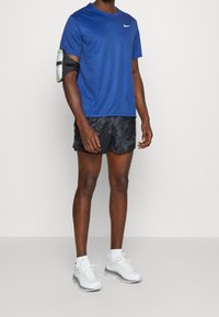 Man står med en blå Nike-t-shirt, svarta mönstrade shorts, vita sneakers och en svart armrem med en transparent ficka.