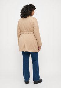 Beige belted frakk med en glatt tekstur, lange ermer og ventilert bak, parret med blå flagrende jeans og svarte sko.