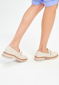 Chaussures en cuir verni beige avec un talon bas, un bout rond, un détail de nœud décoratif et une semelle en caoutchouc texturée.