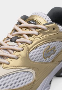 Κοντινό πλάνο λευκού και χρυσού αθλητικού παπουτσιού με υφασμάτινο δίχτυ, επενδεδυμένη γλώσσα, λευκά κορδόνια και το λογότυπο του κροκοδείλου Lacoste στην πλευρά.