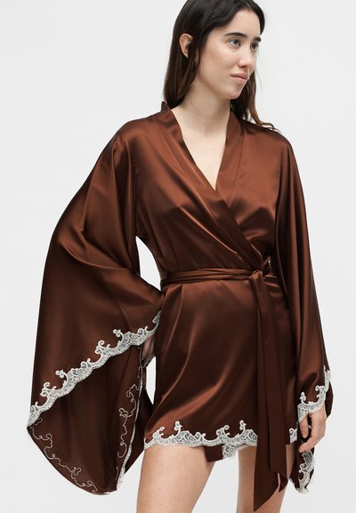Γυναίκα που φορά ένα καφέ σατέν κιμονό ρόμπα με φαρδιά μανίκια και λευκή δαντέλα περιμετρικά, δεμένη στη μέση με μια ζώνη στο ίδιο χρώμα.