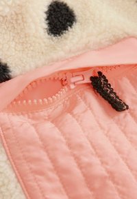 Primer plano de una tela acolchada rosa con una cremallera rosa a juego parcialmente abierta, con un tirador de cremallera negro tejido a crochet y forro polar beige con manchas negras.
