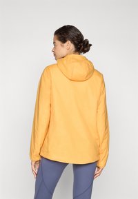 Veste à capuche jaune avec une texture lisse et des manches longues, offrant une coupe décontractée et une capuche ajustable. Portée par-dessus des leggings bleus.