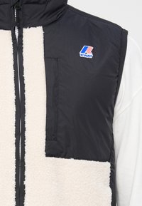 Gilet sans manches noir avec un panneau avant en polaire blanche, arborant un logo K-Way et une fermeture éclair. Texture douce et matériaux contrastés.