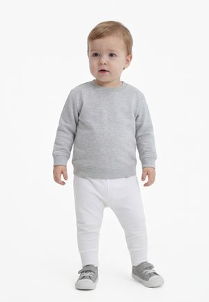 Peuter staand, gekleed in een lichtgrijze sweatshirt, witte broek en grijze sneakers, tegen een effen witte achtergrond.