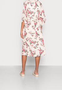 Kwiatowa sukienka midi z krótkimi rękawami, z kremowym tłem i różowymi kwiatami, związana w talii. Noszona z casualowymi beżowymi sandałami.