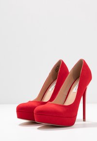 Chaussures à talons hauts en cuir verni rouge avec un bout arrondi et un design plateforme, présentant une texture lisse et un talon aiguille étroit.