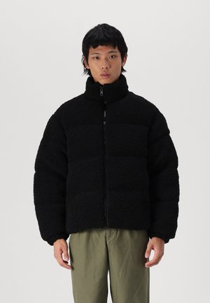 Veste noire en velours, zippée, avec col haut, sections matelassées et coupe décontractée. Elle présente des poches latérales et un tissu texturé et pelucheux.