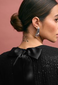 Black sequined garment with a satin bow at the back; model wearing shiny silver earrings. Background is a soft pink.