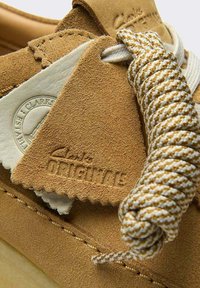 Zamszowy but w kolorze koniakowym o fakturowanej powierzchni, z jasnobarwnym sznurowadłem i metką z napisem „Clarks Originals” wytłoczonym na brązowo.