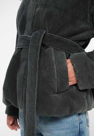 Tumehall korkpa jacket vööga, millel on tekstureeritud pind, küljetaskud ja laiad varrukad, kombineeritud helesiniste teksadega.