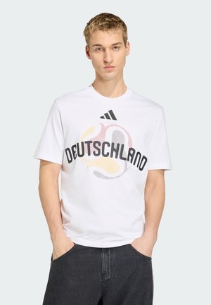 Giovane uomo che indossa una t-shirt bianca Adidas con la scritta "Deutschland" e un design astratto di calcio, mani in tasca, sfondo neutro.
