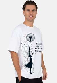 Wit T-shirt met een zwarte silhouet van een vrouw met een paardebloem, met de tekst "Verander jezelf om de wereld te veranderen." Korte mouwen.