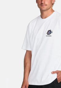 T-shirt en coton blanc avec un col rond, des manches courtes et un design graphique coloré comprenant un visage et un logo sur la poitrine gauche.