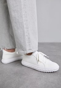 Baskets blanches en tissu flexible, avec un design texturé, un bout rond et une semelle à motifs, portées avec un pantalon gris clair.