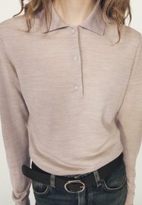 Beżowa koszulka polo z długim rękawem, kołnierzykiem, trzema guzikami z przodu i miękką fakturą, zestawiona z czarnym paskiem i dżinsami.