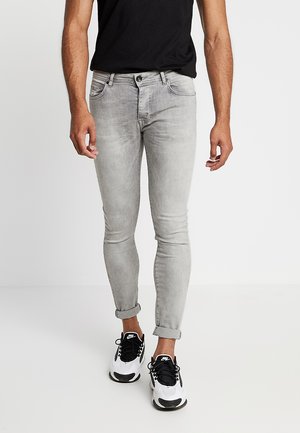 Grijze stretchjeans met een slim fit, voorzien van opgerolde mouwen en voorzakken. Gecombineerd met zwarte sneakers en een zwart T-shirt.