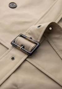 Beżowa tkanina trencza z czarną prostokątną klamrą, gładka tekstura, widoczne szwy oraz metalowe detale.