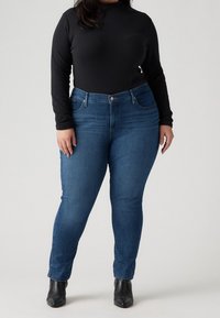 Jeans ajustados en denim azul oscuro con una cintura de altura media, cinco bolsillos y una textura suave, combinados con una blusa negra de manga larga ajustada.