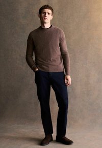 Brauner Strickpullover mit rundem Ausschnitt, kombiniert mit dunkler Denim-Hose und braunen Stiefeletten. Vor einem strukturierten, neutralen Hintergrund.
