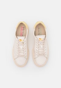 Un par de zapatillas blancas de caña baja con suelas beige, cordones beige claros y pestañas en el talón amarillas, vistas desde arriba sobre un fondo blanco.