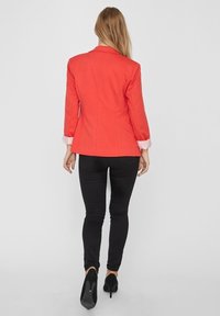 Rode op maat gemaakte blazer met lange mouwen en gestreepte manchetdetails, gecombineerd met zwarte slim-fit broek en zwarte hoge hakken.