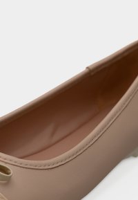 Κοντινό πλάνο μιας μπεζ δερμάτινης μπαλαρίνας που δείχνει ομαλή υφή, ραφές και ένα μικρό διακοσμητικό φιόγκο κοντά στη φτέρνα.