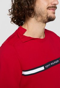 Kiharakateid omav mees kannab punast Tommy Hilfigeri särk, millel on must ja valge triip ning kaelus on kaela lähedal lahti.