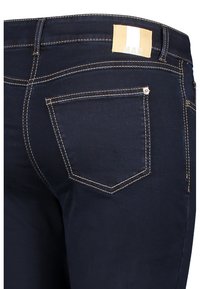 Vaqueros de mezclilla azul oscuro con costuras en crema en contraste, que cuentan con un bolsillo trasero con botón y una etiqueta de la marca en la cinturilla.