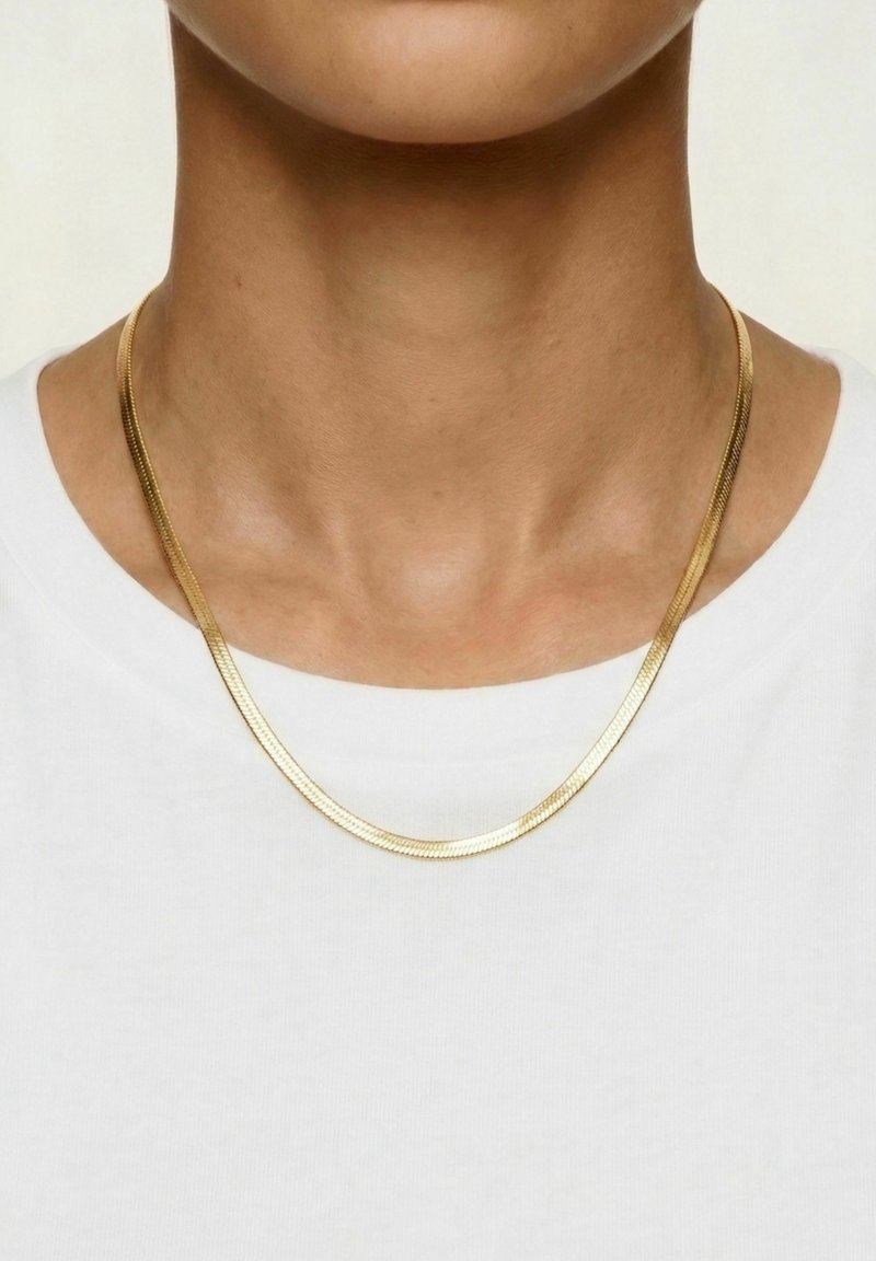 Gros plan d'une personne portant une fine chaîne plate en or par-dessus une chemise blanche unie sur un fond neutre.