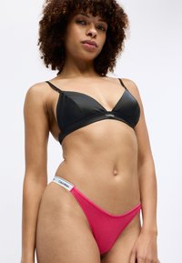 Svart triangelformad bralette och rosa tanga, tillverkade av ett slätt tyg. Braletten har justerbara remmar, medan tangat har en midjeband med logotyp.