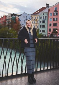 Giubbotto puffer nero sopra un vestito in maglia a righe grigie, abbinato a stivali neri alla caviglia. Sullo sfondo ci sono edifici dai colori pastello vicino all'acqua.
