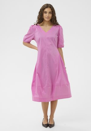 Göndör hajú nő élénk rózsaszín midi ruhában, puffos ujjakkal és fekete hegyes orrú magassarkú cipőben áll fehér háttér előtt.