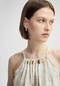 Jasnozłoty halter top z plisami, ozdobiony kamieniami w różnych odcieniach wzdłuż dekoltu; tkanina ma subtelny wzór.