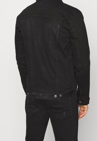 Svart jeansjacka med en figursydd design, knappdetaljer vid midjan och strukturerad tyg. Avslutad med krage och sömmar på baksidan.