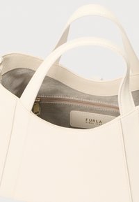 Balta ādas rokassoma ar diviem rokturiem, teksturētu ārpusi un mīkstu auduma oderi ar rāvējslēdzēja nodalījumu un zīmola uzlīmi.