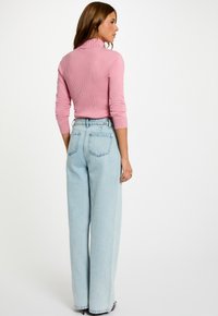 Donna con lunghi capelli castani che indossa un maglione rosa a coste e jeans a vita alta, larghi e di colore blu chiaro, in piedi contro uno sfondo bianco.