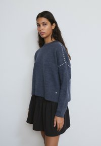 Mujer con cabello largo y oscuro, vestida con un suéter azul oscuro con tachuelas plateadas en las mangas y una falda negra, de pie frente a un fondo blanco liso.