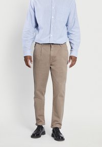 Beige katoenen broeken met een recht model, voorplooien en zijzakken, gecombineerd met een blauw-wit gestreept overhemd met knopen en zwarte schoenen.